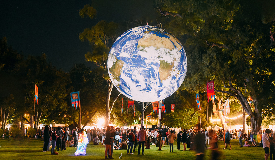 A lit up globe hovers above people walking on grass; trees and colourful flags are visible in the background.