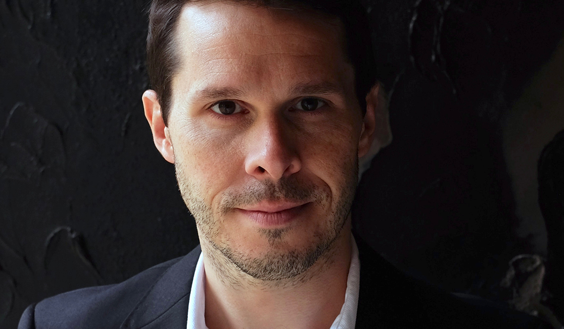 David Greco has dark hair and a small amount of dark and grey stubble. He is wearing a suit and looking at the camera with a half-smile.