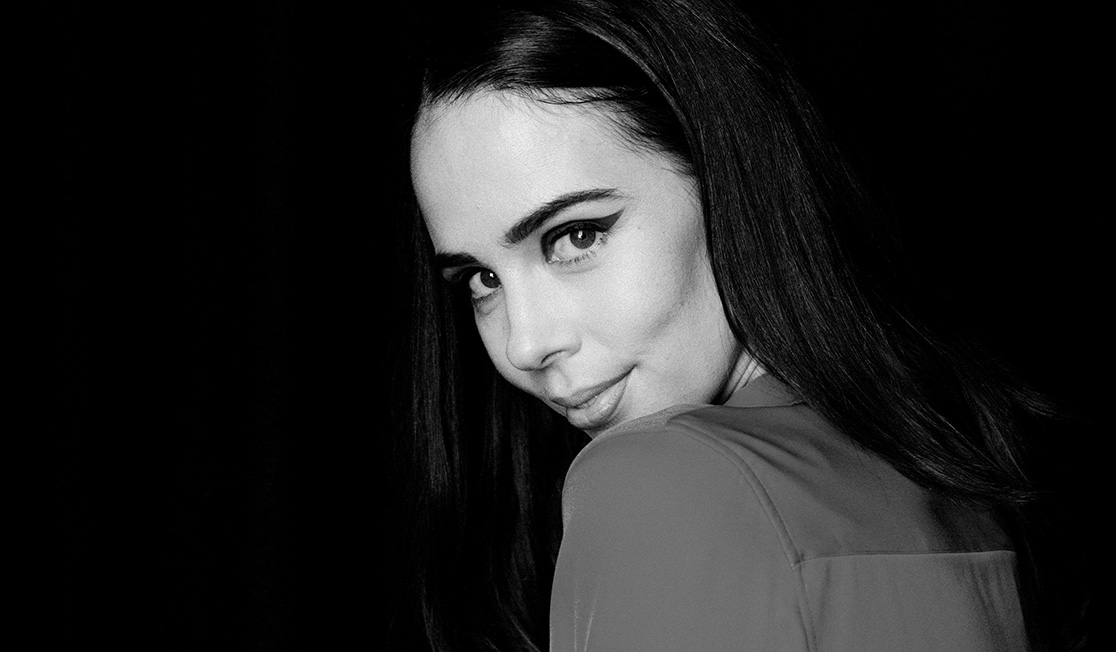 A black and white image of a woman with long dark hair looking over her shoulder. She is looking at the camera with a coy expression.