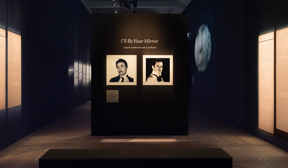 Two black and white photos hang on a black wall. One is a photo of Laurie Anderson and the other is a photo of Loud Reed - both of them wear ties and mustaches and look at the camera with slightly raised brows and serious expressions. Above the photos are the words "I'll be your mirror".