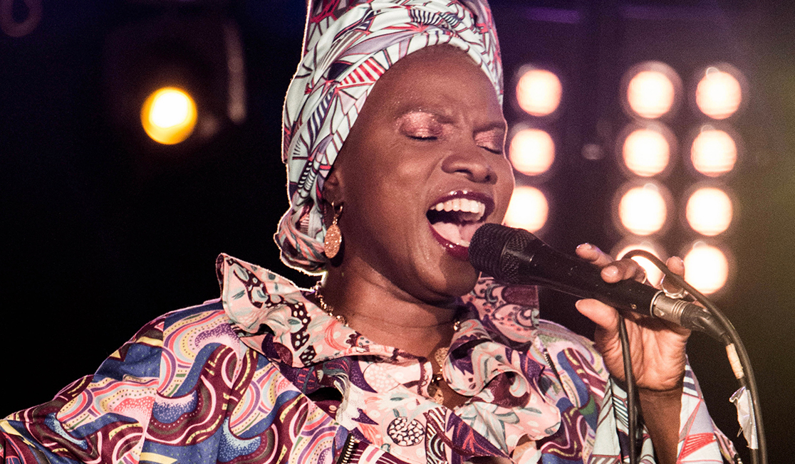 A woman wearing a head wrap and shirt made from patterned material in maroon, cream and blue sings into a microphone with her eyes closed.
